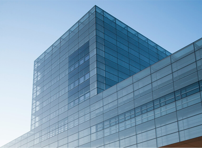 Glass-walled modern building with a blue tint, featuring a combination of rectangular shapes against a clear sky.