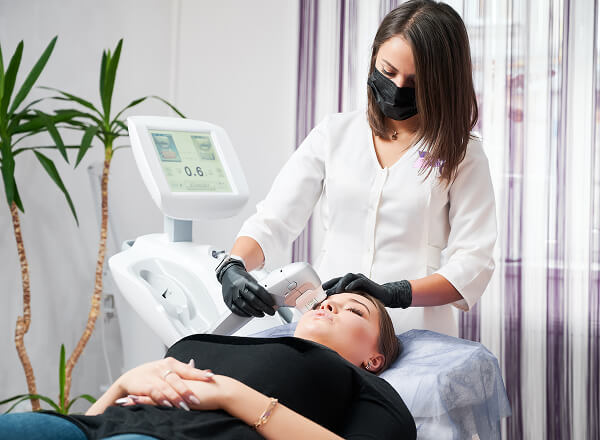 A cosmetologist wearing gloves and a face mask performs a facial laser treatment on a woman lying on a clinic bed.