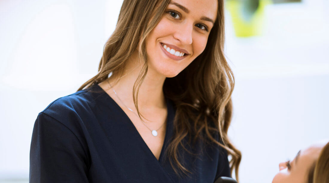 Woman in navy scrubs smiling at the camera, standing indoors with another person partially visible in the foreground.