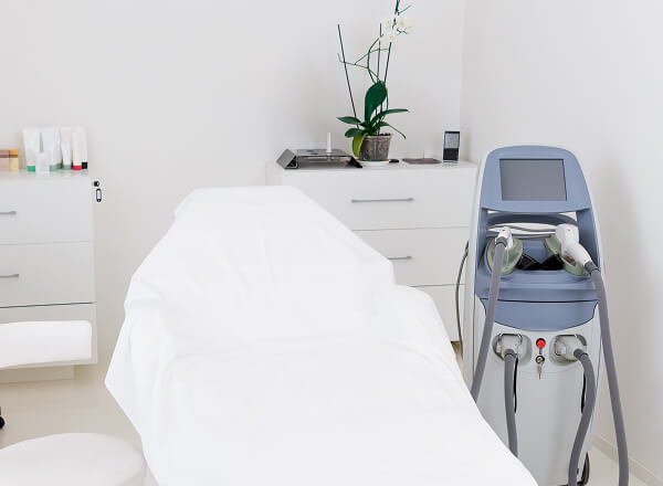 A treatment room with a covered examination bed, a medical device with a screen and handpieces, white cabinets, and a potted plant on the counter.
