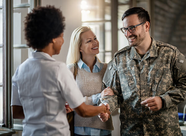A smiling man in military uniform shakes hands with a woman in a white top while another woman stands beside him, all appearing friendly and happy indoors.