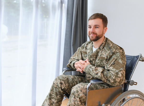Man in military uniform sitting in a wheelchair near a window with sheer curtains, looking outside and smiling.
