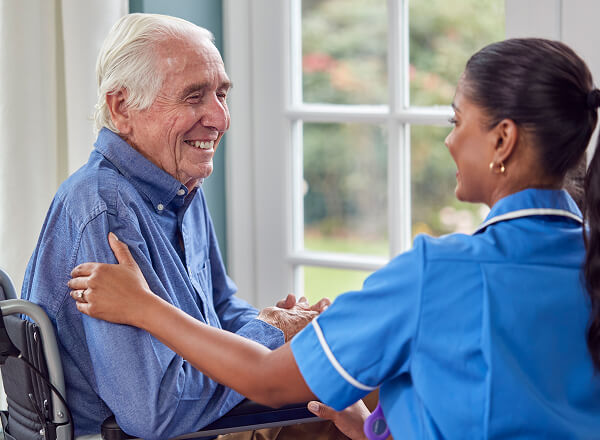 Elderly man in a wheelchair smiles while a nurse in blue uniform gently holds his shoulder in a bright room.