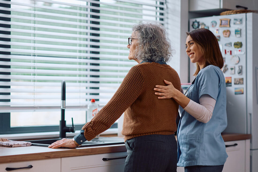 A caregiver in scrubs stands beside and supports an elderly woman who is looking out the kitchen window.