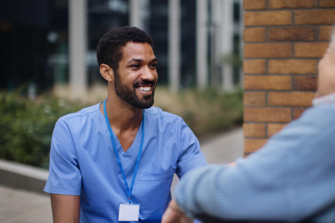 A healthcare worker in blue scrubs and an ID badge smiles while talking to another person outdoors near a brick wall.