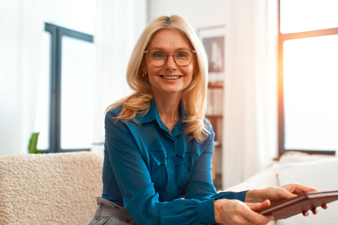 A woman with blonde hair and glasses, wearing a blue blouse, sits on a sofa indoors, smiling and holding a tablet.