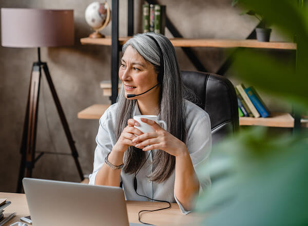Woman with long gray hair sits at a desk, wearing a headset and holding a mug, looking to the side. A laptop, books, and houseplant are visible in the background.