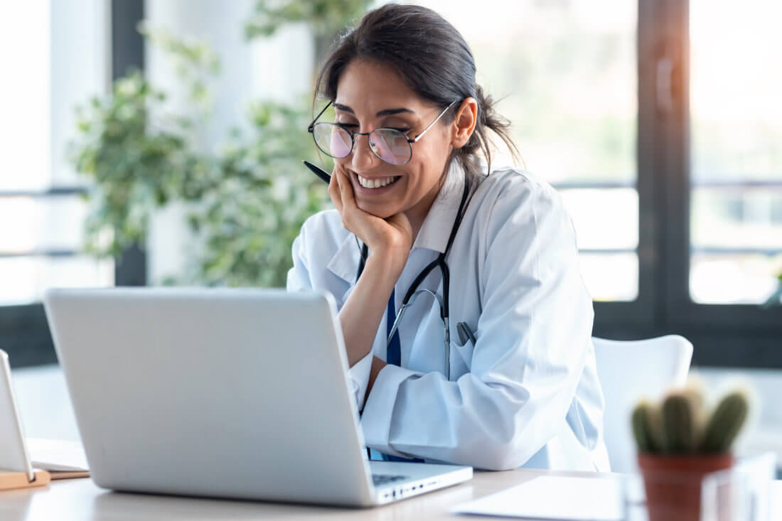 Female doctor wearing a white coat and stethoscope smiles while looking at a laptop in a bright office setting.