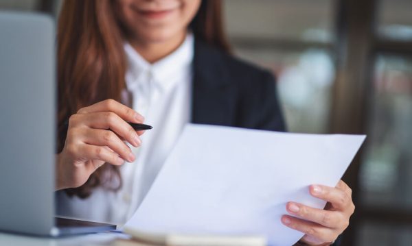 A woman in business attire sits at a desk, holding a pen in one hand and a sheet of paper in the other, with a laptop nearby.