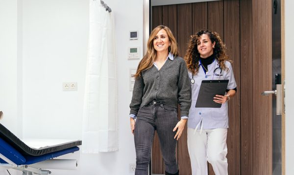 A smiling patient walks out of a medical examination room accompanied by a healthcare professional holding a clipboard.
