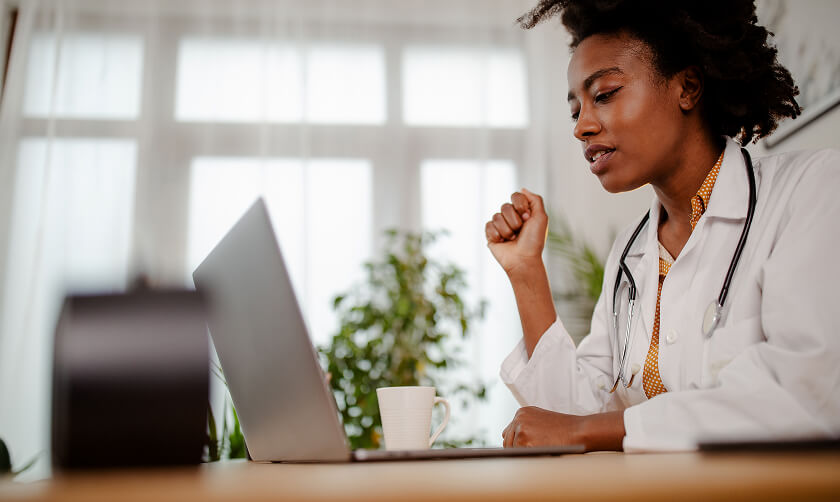 A doctor wearing a white coat and stethoscope sits at a desk, looking at a laptop screen with a coffee cup nearby.