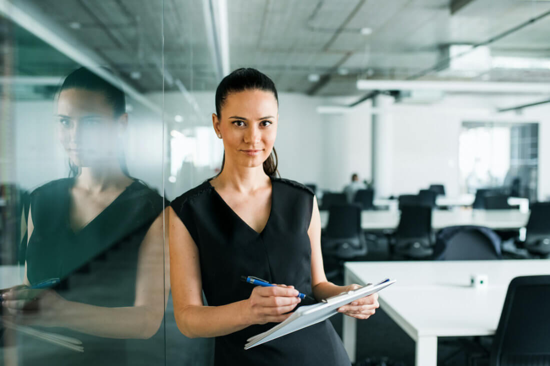 A woman stands in an office holding a clipboard and pen, looking at the camera. She is next to a glass wall with empty desks and chairs in the background.