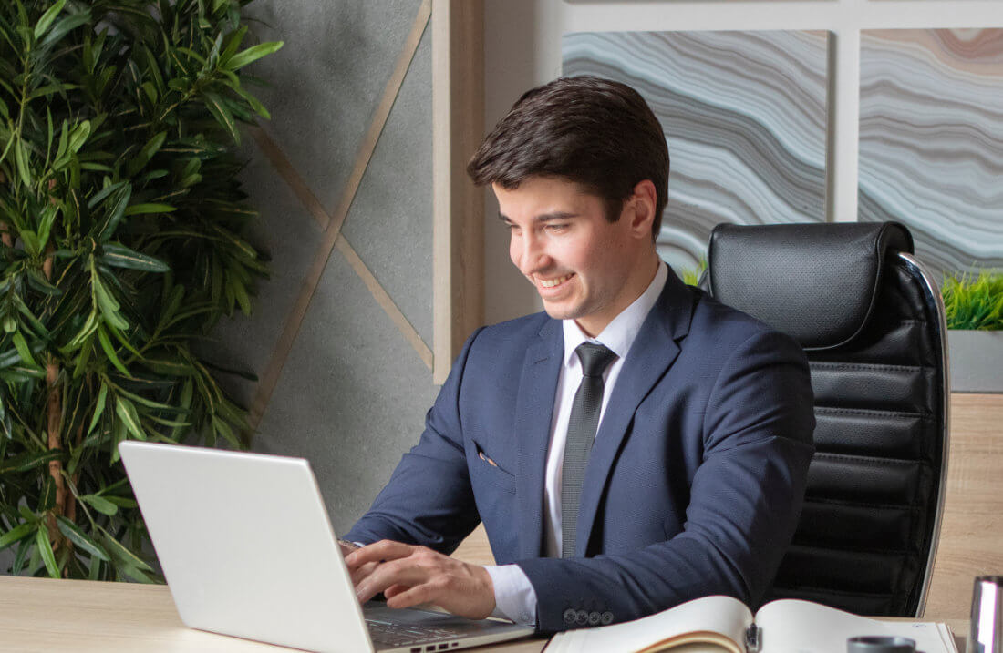 A man in a suit sits at a desk, smiling while typing on a laptop. An open notebook and office decor are visible in the background.