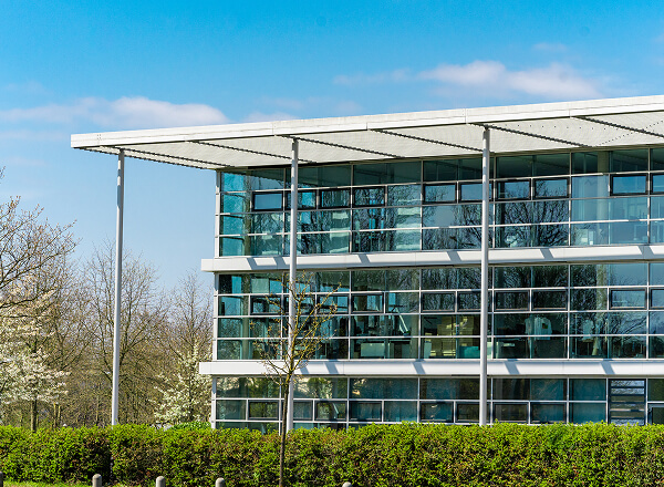 Modern glass office building with three visible floors, surrounded by green hedges and trees, under a clear blue sky.