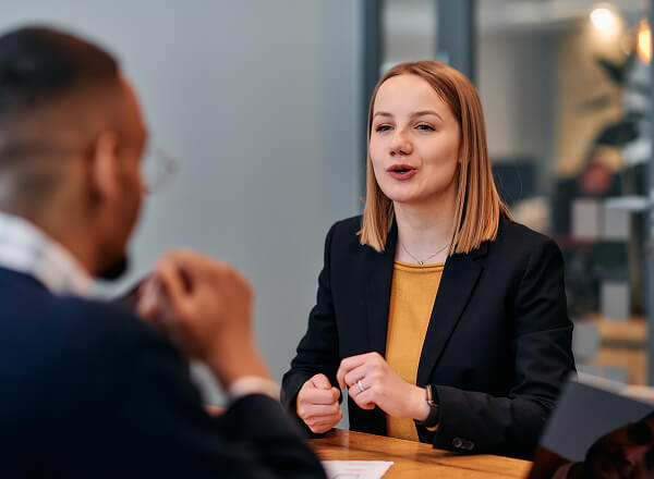 Two people sit across from each other at a table in a professional setting, engaged in conversation. The woman is speaking and gesturing with her hands.