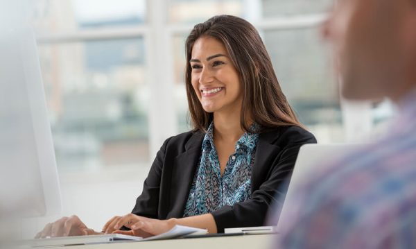 A woman sits at a desk, smiling at a computer screen, with papers and a laptop in front of her in a bright office setting.