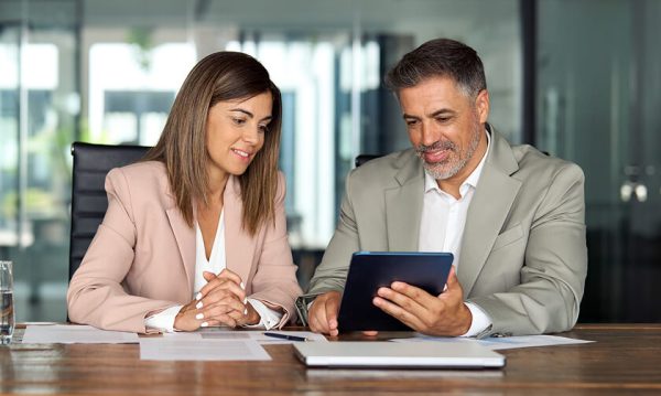 Two business professionals sit at a table, reviewing information on a tablet, with papers and a glass of water nearby in a modern office setting.