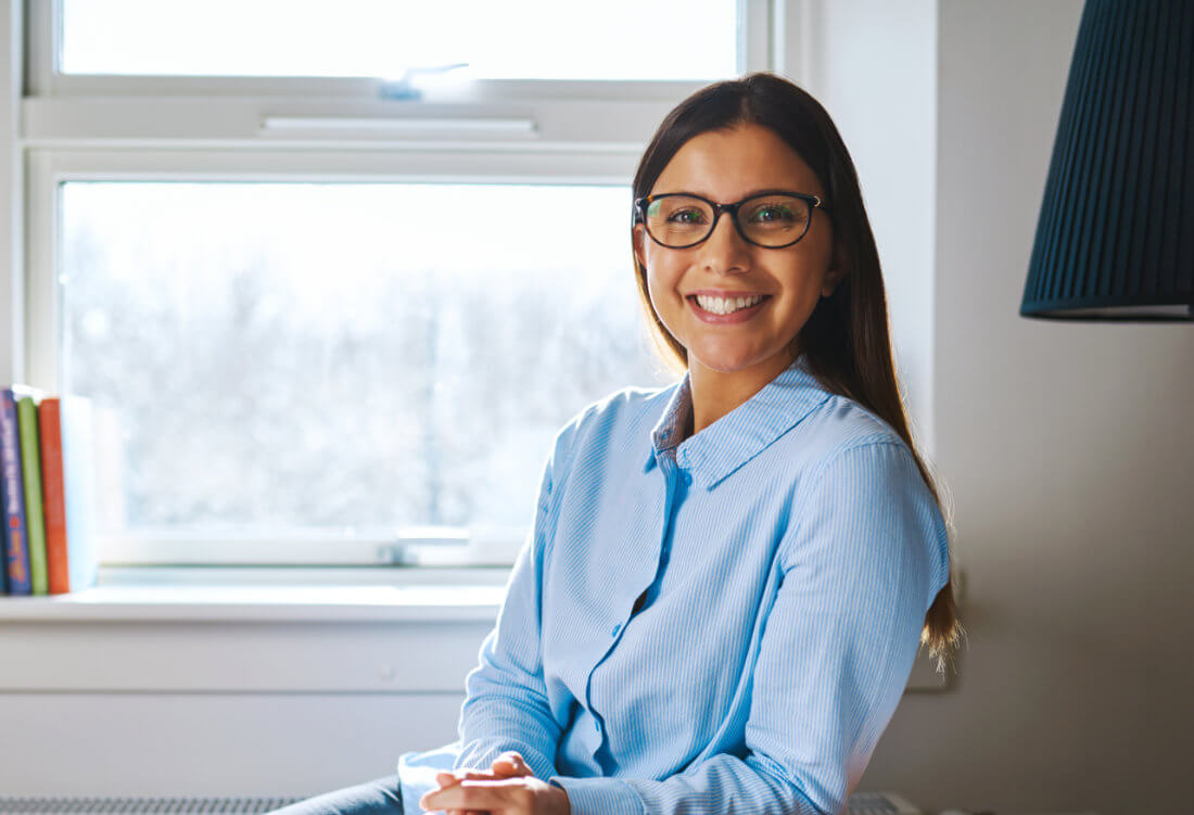 A woman wearing glasses and a light blue shirt sits indoors by a window, smiling at the camera. A stack of books is visible on the windowsill in the background.
