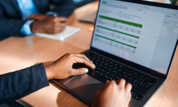 A person types on a laptop displaying a spreadsheet, while another person writes notes on paper at a desk.