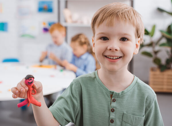 Smiling boy holds a pink clay figure in the foreground; two children work at a table in the blurred background.