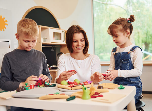 A woman sits at a table with a young boy and girl, helping them play with toy food in a bright, child-friendly classroom.