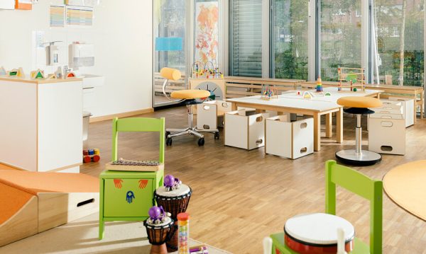 A brightly lit classroom with wooden floors, child-sized tables and chairs, green stools, educational toys, and large windows letting in natural light.