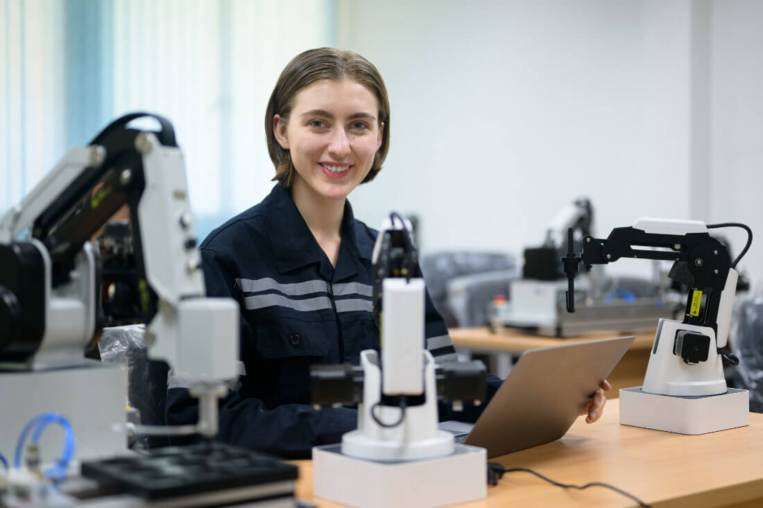 Person in a dark uniform sits at a desk with a laptop, surrounded by robotic arms in a modern, well-lit laboratory or classroom.