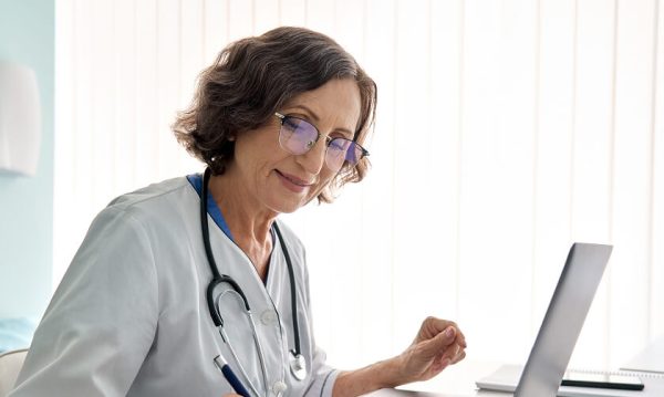 An older female doctor wearing glasses and a white coat sits at a desk with a laptop, writing notes with a pen. A stethoscope hangs around her neck.