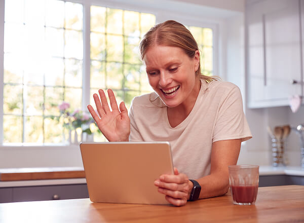 Woman sitting at a kitchen table waving at a tablet during a video call, with a glass of juice beside her.