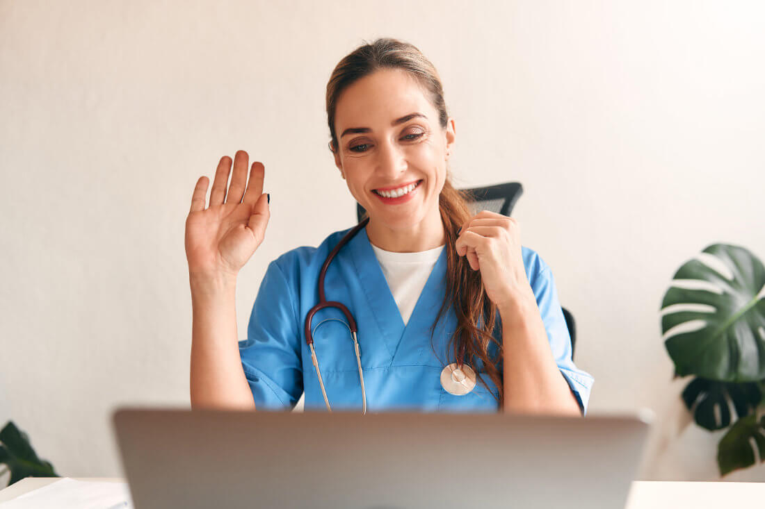 A healthcare professional in blue scrubs and a stethoscope smiles and waves at a laptop during a video call.