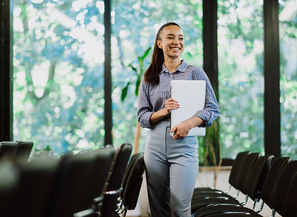 Woman standing in a conference room holding a laptop, smiling, with rows of empty chairs and large windows showing greenery in the background.