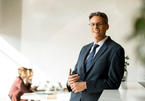 A man in a business suit stands and smiles in an office, with two people working at desks in the background.