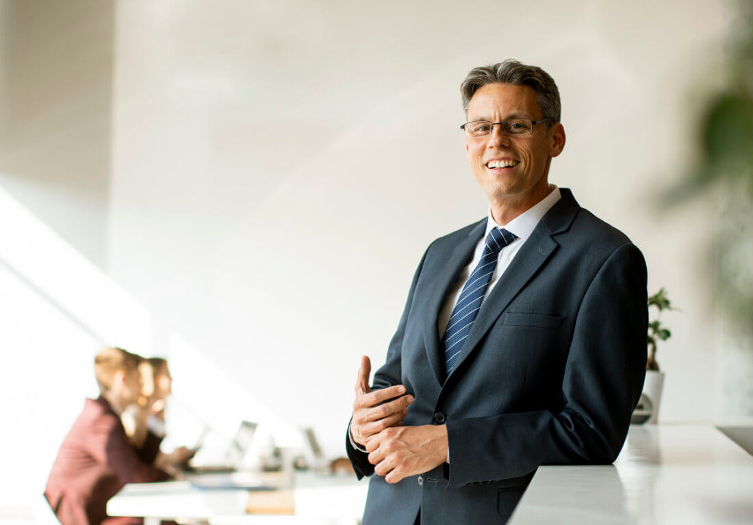 A man in a business suit stands and smiles in an office, with two people working at desks in the background.