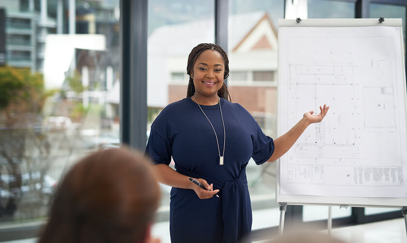 A woman stands next to a flip chart with architectural plans, gesturing as she presents to a group in a modern office setting.