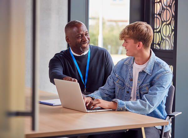 An older man and a younger man sit at a table with a laptop, having a conversation. The older man is smiling and pointing at the screen.