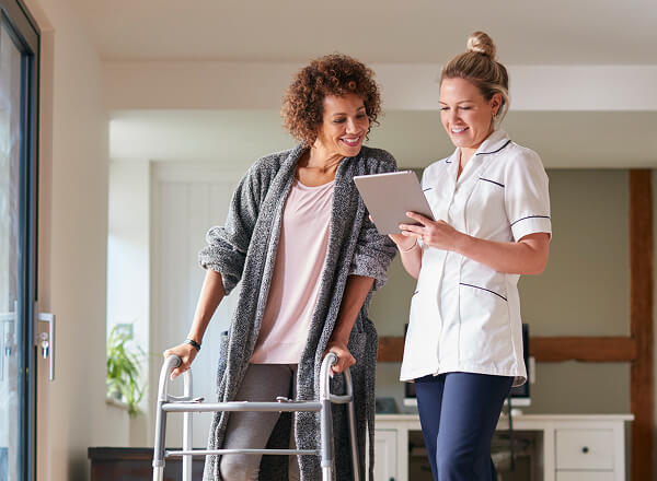 A woman using a walker stands next to a healthcare professional who is showing her information on a tablet.