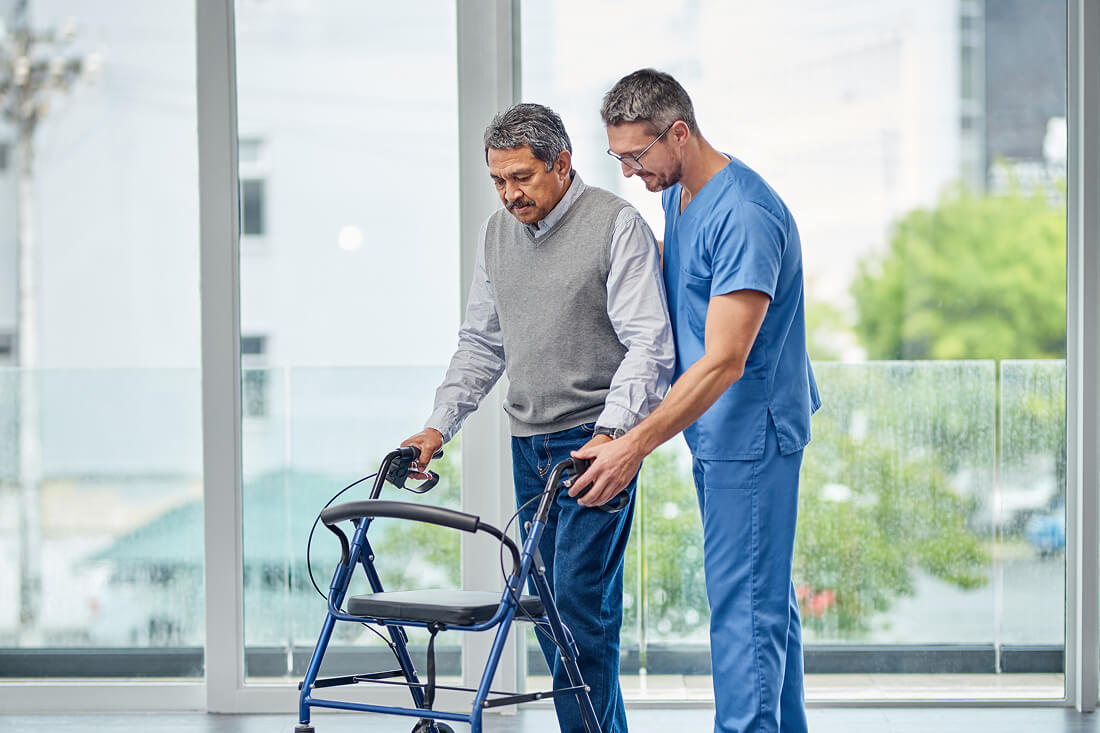 A healthcare professional assists an older man using a walker in a bright, modern indoor setting with large windows.
