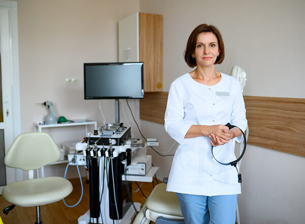 A female medical professional stands in a clinic room, holding an endoscope, with examination equipment and a monitor in the background.