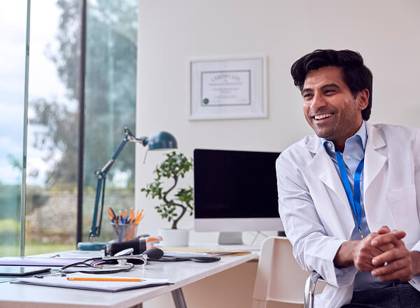 A doctor in a white coat sits and smiles in a modern office with a computer, stethoscope, and paperwork on the desk. A certificate hangs on the wall in the background.
