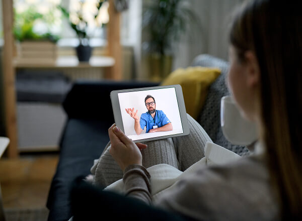 A woman sits on a couch holding a tablet, having a video call with a male doctor who is wearing blue scrubs and glasses.