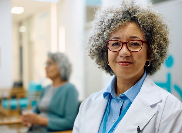A woman wearing a white lab coat and glasses smiles at the camera, with another person seated and blurred in the background.