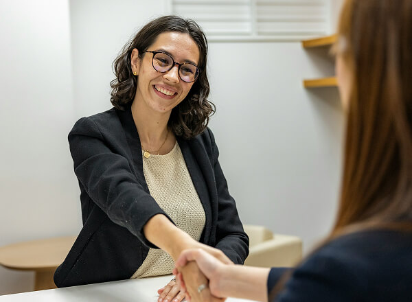 A woman in business attire smiles while shaking hands with another person across a desk in a modern office setting.