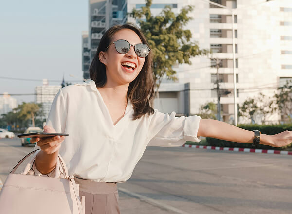 A woman wearing sunglasses is smiling and hailing a taxi on a city street while holding her phone and a handbag.