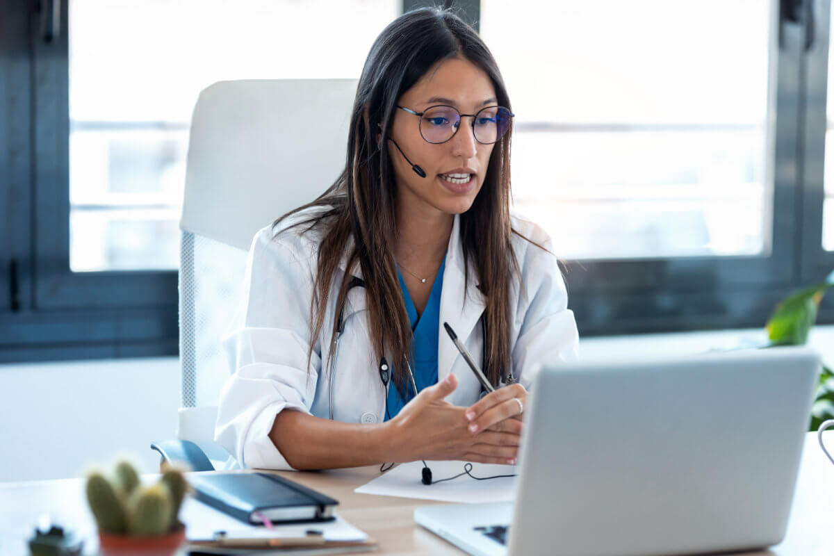 A doctor wearing a headset and white coat sits at a desk, speaking during a video call on a laptop, with documents and a stethoscope in view.