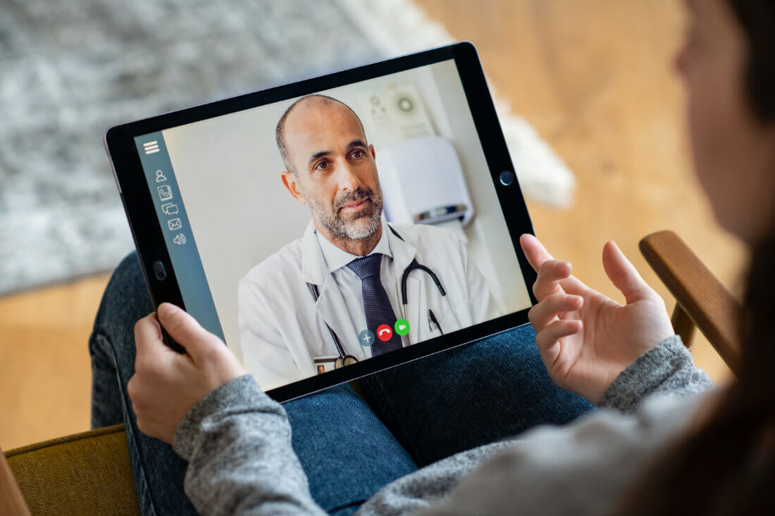 A person holds a tablet showing a video call with a doctor in a white coat and stethoscope.