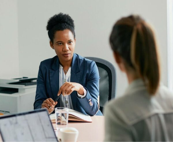 Two professional women in an office, one african descent explaining something to the other, who is facing away from the camera.