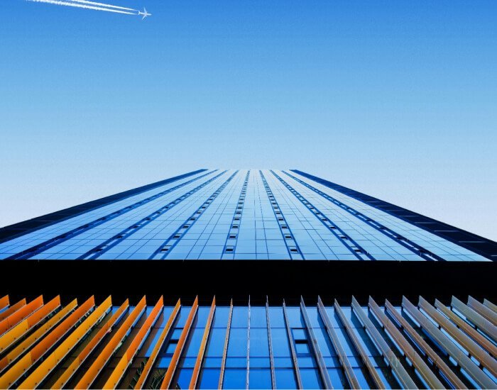 Looking up the façade of a modern skyscraper with blue and orange glass panels, against a clear sky with a distant airplane leaving a white trail.