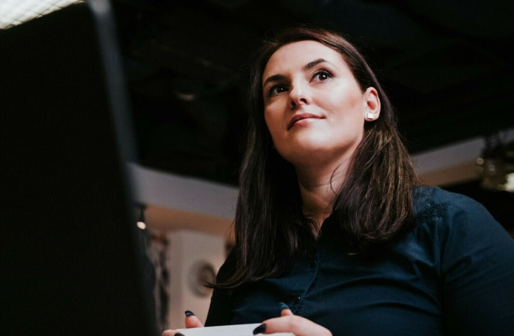 A woman in HCPA focused on her computer screen in a dimly lit room, holding a pen and appearing thoughtful.
