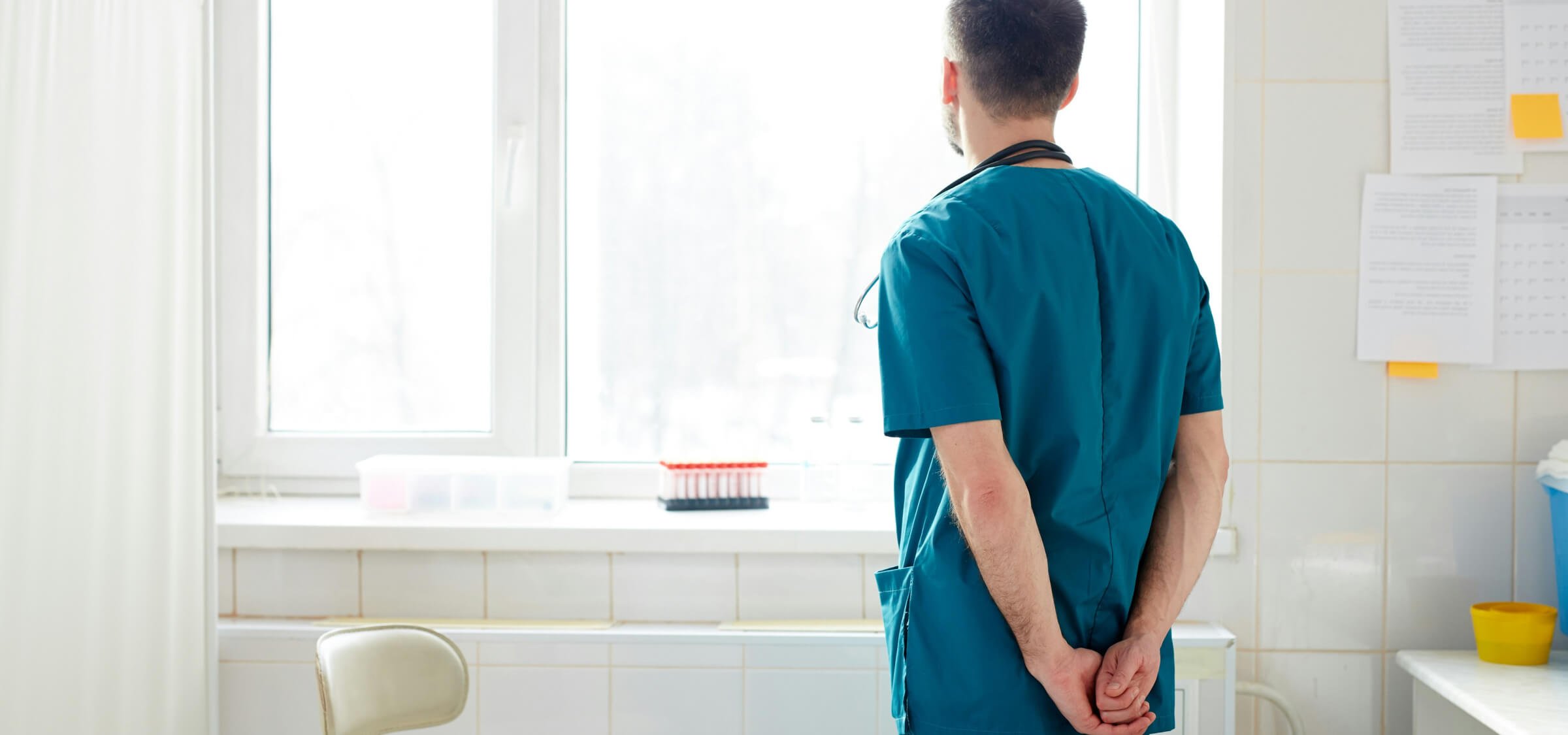 A male healthcare worker in scrubs standing by a window in a brightly lit room, looking outside, with his hands clasped behind his back.
