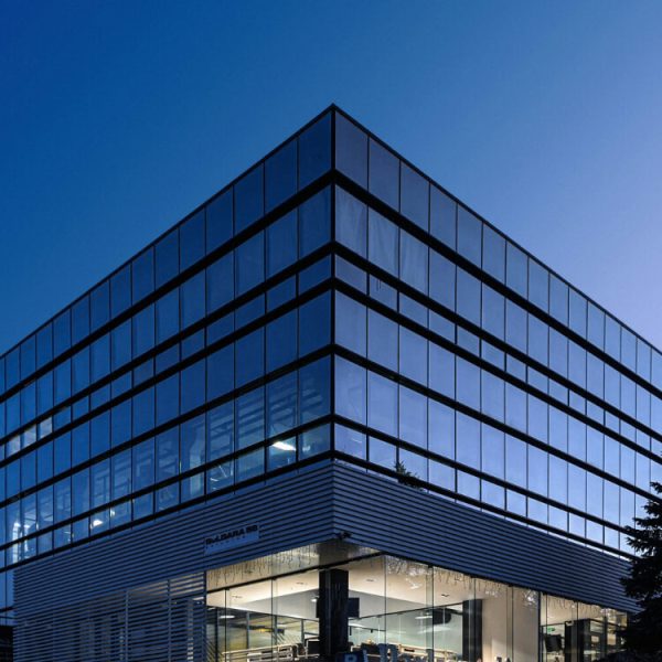 A modern glass office building with illuminated interior lights, photographed at dusk against a clear blue sky.